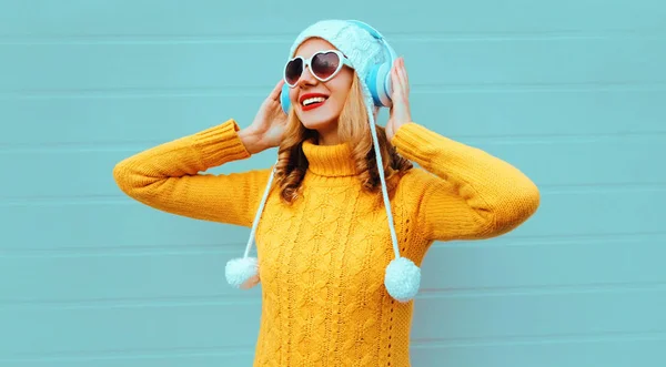 Winter portrait happy smiling young woman in wireless headphones listening to music wearing yellow knitted sweater and white hat with pom pom, heart shaped sunglasses on blue wall background