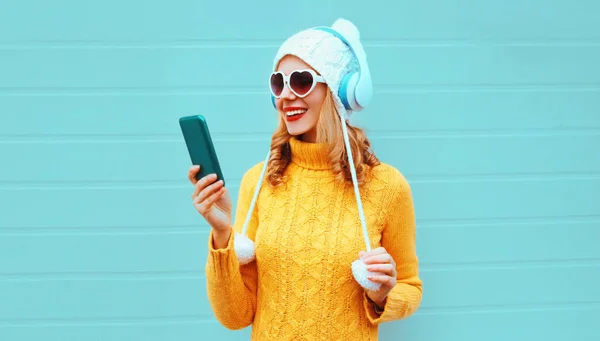 Winter portrait happy smiling woman holding phone in wireless headphones listening to music wearing yellow knitted sweater, white hat with pom pom, heart shaped sunglasses on blue wall background