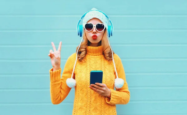 Χειμερινό πορτρέτο δροσερό γυναίκα με τηλέφωνο σε ακουστικά ακούγοντας μουσική που δείχνει την ειρήνη χέρι σημάδι φυσώντας κόκκινα χείλη φορώντας κίτρινο πλεκτό πουλόβερ, λευκό καπέλο, γυαλιά ηλίου σε σχήμα καρδιάς σε μπλε φόντο — Φωτογραφία Αρχείου