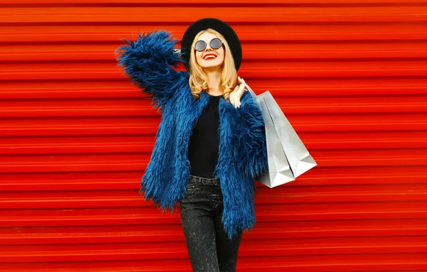 ショッピングバッグとスタイリッシュな笑顔の女性は、赤い壁の背景に青いフェイクファーコート、ラウンド帽子とサングラスを身に着けている新しい成功した購入を楽しんで — ストック写真