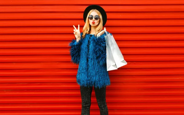 Atrakcyjna młoda kobieta z torbami na zakupy, stylowa modelka pokazująca gest pokoju w niebieskim, sztucznym futrze, okrągłym kapeluszu i okularach przeciwsłonecznych na czerwonym tle ściany — Zdjęcie stockowe
