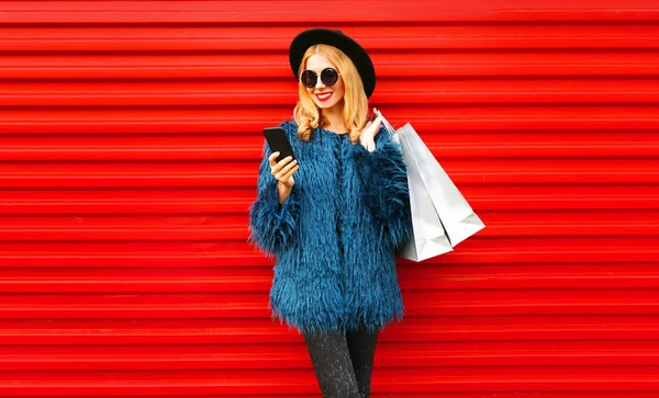 携帯電話やショッピングバッグを身に着けているスタイリッシュな笑顔の女性青いフェイクファーコート、ラウンド帽子と赤い壁の背景にサングラス — ストック写真