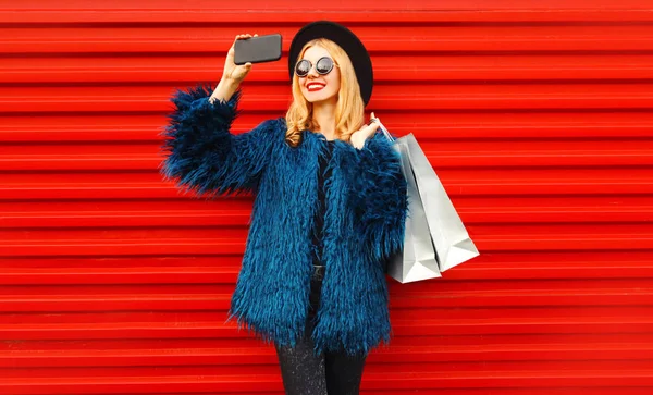 魅力的な若い笑顔の女性がショッピングバッグでスマートフォンで写真を撮る、スタイリッシュな女性モデルは、赤い壁の背景に青いフェイクファーコート、黒丸帽子とサングラスを身に着けている — ストック写真
