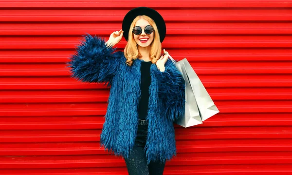 ブルーのフェイクファーコート、黒の丸帽子と赤い壁の背景の上にポーズサングラスを身に着けているショッピングバッグとポートレートスタイリッシュな笑顔女性 — ストック写真