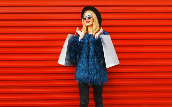 ショッピングバッグを身に着けているスタイリッシュな笑顔の女性青いフェイクファーコート、黒丸帽子と赤い壁の背景にサングラス — ストック写真