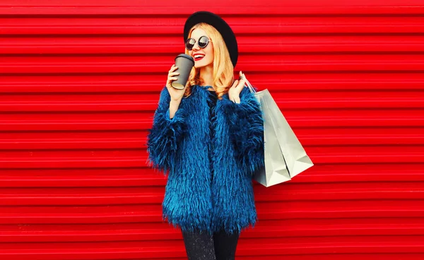 肖像画スタイリッシュな笑顔の女性コーヒーを飲んでショッピングバッグを保持青いフェイクファーコート、黒丸帽子とサングラスを身に着けている赤い壁の背景の上にポーズ — ストック写真