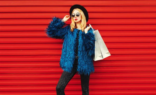 魅力的な女性ポージングでショッピングバッグ、スタイリッシュな女性モデル着用青フェイクファーコート、黒丸帽子とサングラス以上赤壁の背景 — ストック写真