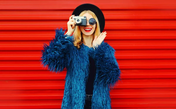 Retro kameralı, mavi kürklü, yuvarlak şapkalı ve güneş gözlüklü, renkli kırmızı duvarlı bir kadın fotoğrafı çekiyor. — Stok fotoğraf