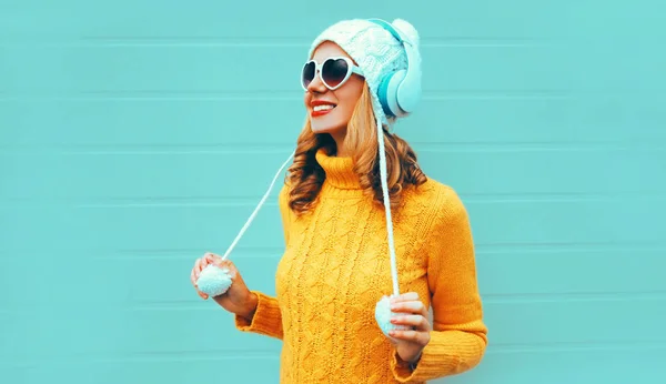 Winter portrait smiling young woman in wireless headphones listening to music wearing yellow knitted sweater and white hat with pom pom, heart shaped sunglasses on blue wall background