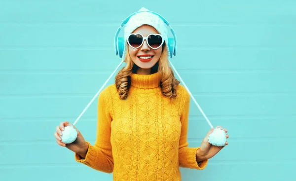 Winter portrait smiling young woman in wireless headphones listening to music wearing yellow knitted sweater and white hat with pom pom, heart shaped sunglasses on blue wall background