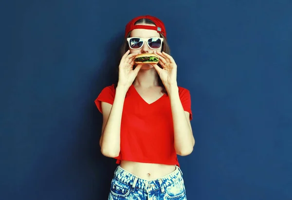 Cool young woman eating burger wearing baseball cap, sunglasses