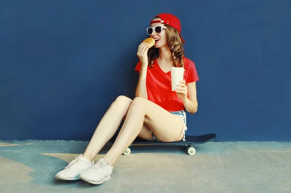Cool young woman with burger and coffee cup sitting on skateboard