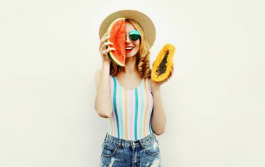 Bir dilim karpuz ve papaya dilimli çekici bir kadın hasır şapka takıyor, güneş gözlüğü takıyor, beyaz arka planda şort giyiyor.
