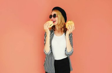 Portre, hamburger yiyen, siyah şapka takan, arka planda güneş gözlüğü takan modern bir genç kadın.