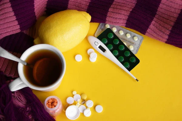 Assorted pharmaceutical medicine pills, tablets and capsules on yellow background. A cup of tea with lemon and a warm scarf. Disease. Maintenance of immunity. Seasonal diseases. Top view. Medicine flat lay