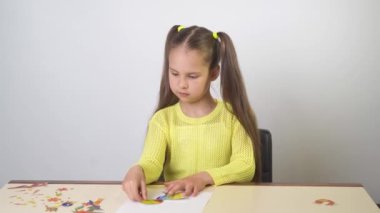 8 yaşındaki bir kız, sarı bir bluz içinde beyaz bir arkaplanda kompozisyon oluşturmak için gülümser ve bir sayfa kağıt keser.