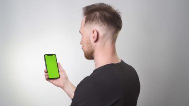 Adamın elinde yeşil arkaplanı olan bir telefon var. Arkası kameraya dönük, siyah tişörtlü beyaz bir arka planda duruyor.