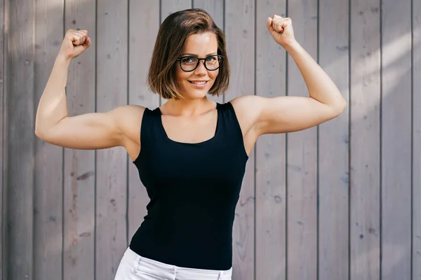 Kısa saçlı güçlü fitness kızı, sıradan giyinmiş, pazılarını ahşap arka planda gösteriyor.