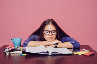 Gözlüklü esmer kız ve mavi gri takım elbiseli kız elinde not defteri, tablet, kalemler, telefon ve mavi bardakla masada oturuyor, pembe arka planda ellerini üzerine koyuyor. Arkaplanda izole edilmiş insanlar