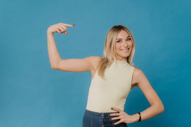 Açık bluzlu ve kot etekli neşeli, aktif ve iyimser beyaz kız kendini göstererek gülümsüyor, mavi arka planda yaptığı işlerden gurur duyuyor..