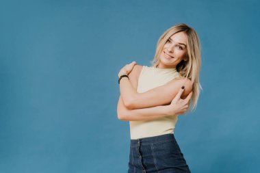 Açık renk bluz ve kot etek giyen hoş bir sarışın, stüdyoda güzel bir gülümsemeyle poz veriyor, kendine sarılıyor, kendine güveniyor, keyfi yerinde, mavi arka planda fotokopi çekiyor. Mutlu insanlar konsepti.