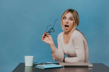 Masada dizüstü bilgisayarlı, günlüklü açık pembe bluzlu çekici bir sarışın, ekonomi haberlerine hayran, ağzı ve gözleri açık, mavi arka planda gözlüğünü tutuyor..