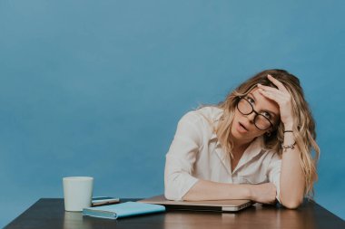 Gözlüklü, beyaz gömlekli, iş yerinde dizüstü bilgisayarla oturan yorgun ve karantina sırasında mavi arka planda çalışmaktan sıkılmış bir sarışın. Coronavirüs salgını konsepti.