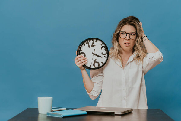 Tired blonde in glasses wearing white shirt sitting at her working place with laptop, deadline is coming, bored working at home during quarantine over blue backdrop. Coronavirus pandemic concept.