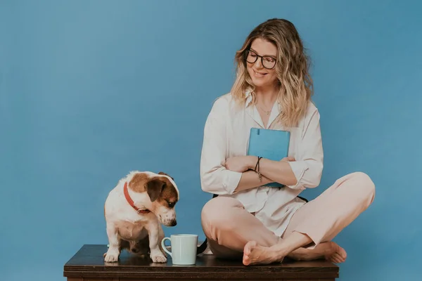 Gözlüklü, beyaz gömlekli, açık pembe pantolonlu sevimli bir sarışındı. Köpek yavrusu Jack Russell 'la aynı masada oturuyordu. Ona şefkatle bakıyordu. Karantina sırasında evde kal. Covid-19 konsepti.