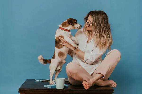 Gözlüklü, beyaz gömlekli, açık pembe pantolonlu sevimli bir sarışın. Köpek yavrusu Jack Russell 'la bir masada oturuyor ve onu öpüyor. Karantina sırasında evde kal. Covid-19 konsepti.