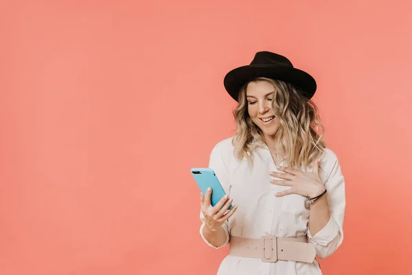 Happy curly blonde in a hat with a brim, white shirt and light pink belt, holding her smartphone, broad smiling, happy to received message, over pink background with copy space. Happy people concept