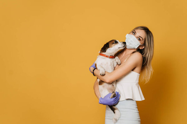 Charming blonde in surgical mask and protective gloves, dressed in a light blouse and skirt hugs her dog, take care about friends during quarantine because of coronavirus pandemic. Covid-19 concept.