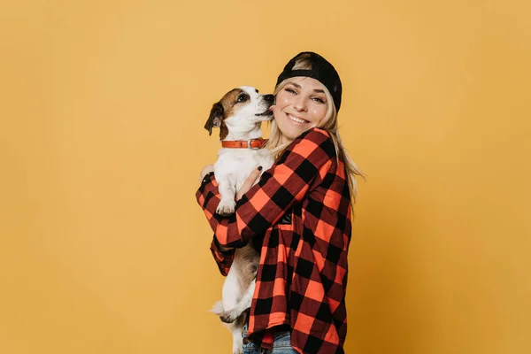 Beyzbol şapkalı genç bir sarışın, gömleğini ve kotunu kontrol etmiş, sırıtan bir kadın, sarı arka planda yanağını yalayan köpeği Jack Russel 'a sarılıyor. Yaşam tarzı ve gençlik kavramı.