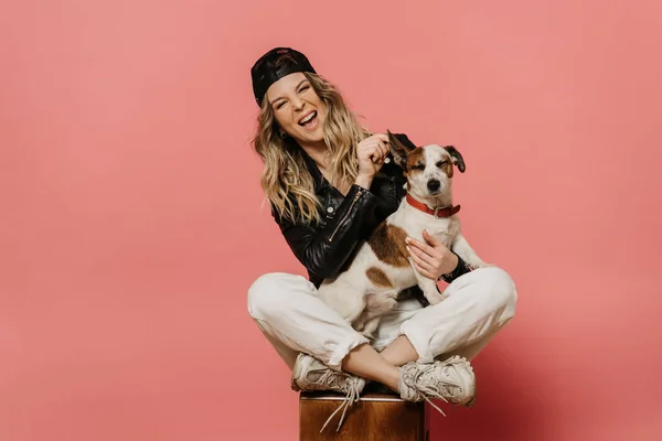 Siyah deri ceket, beyaz kot pantolon ve köpeğiyle oturan güzel sarışın kız. Koronavirüs salgınının bitmesini bekliyor. Pembe renkte, kopyalama alanı ile izole edilmiş. Covid-19 konsepti.