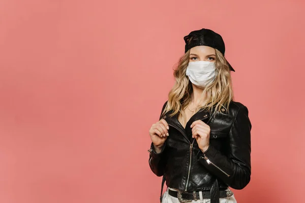 Cerrahi maske takan, siyah deri ceket, beyaz kot pantolon ve şapka giyen sarışın genç ve güzel bir kız. Koronavirüs salgınının bitmesini bekliyor. Pembe renkte, kopyalama alanı ile izole edilmiş. Covid-19 konsepti.