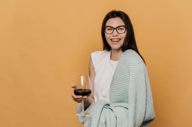 Beyaz bir tişört giymiş, elinde pastel bir ekoseyle sarılı, elinde bir bardak şarapla gülen, sarı arka planda duran ve fotokopi çeken neşeli bir esmer. Evde kal, dikkatli ol..