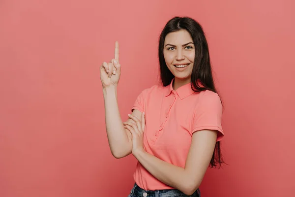 Pembe tişört ve kot pantolon giymiş neşeli, genç esmer kız, geniş bir gülümseme içinde, memnun görünüyor, pembe arka planda işaret parmağıyla yukarıya bakıyor. Satışlar ve indirim.
