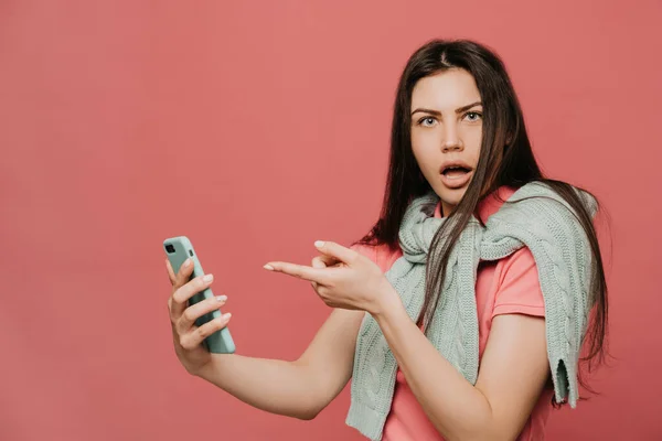 Pembe tişört ve pastel turkuaz süveter giymiş genç bir esmer, boynuna bağlanmış, akıllı telefonunu tutuyor ve pembe arka planda onu işaret ediyor.. 