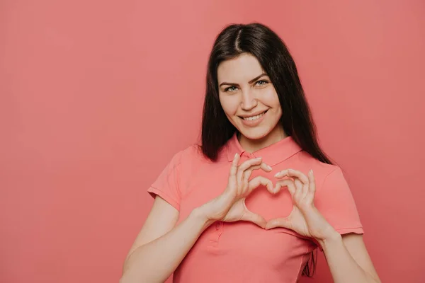 Esmer saçlı çekici bir kadının portresi. Elleriyle kalbi şekillendiriyor ve kameraya dostça gülümsüyor. Sevgi ve özenin sembolü. Pembe arka planda kapalı stüdyo çekimleri
