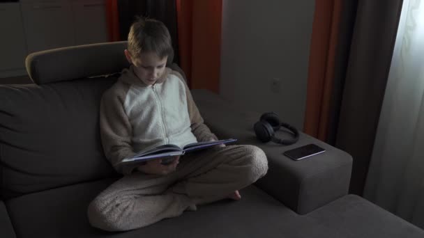 Teen boy reads a book sitting on the couch. He closes the book and rests. quarantine 2020 — Stock Video