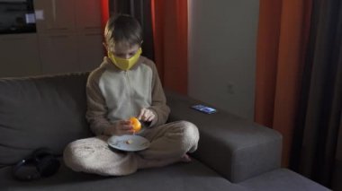 Koruyucu maskeli genç bir adam kanepede otururken mandalina soyar. Coronavirüs salgını 2020.