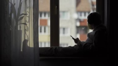Koruyucu maskeli çocuk bir pencere pervazında oturuyor ve elinde telefon var. Karatnin.