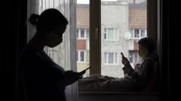 Mulher e adolescente segurando smartphone. ambos em máscaras protetoras em frente à janela. Quarentena 2020 . — Vídeo de Stock