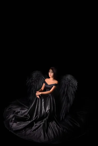 Angel-devil model in black long dress with wings sitting in studio. Black background, — Stock Photo, Image