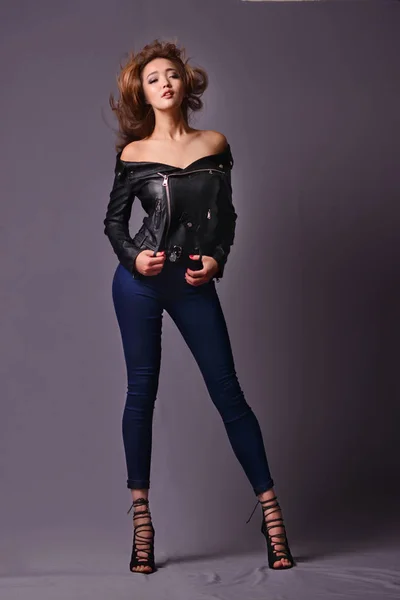 Model with curly hair in a leather jacket, jeans and high heels posing in studio — Stock Photo, Image