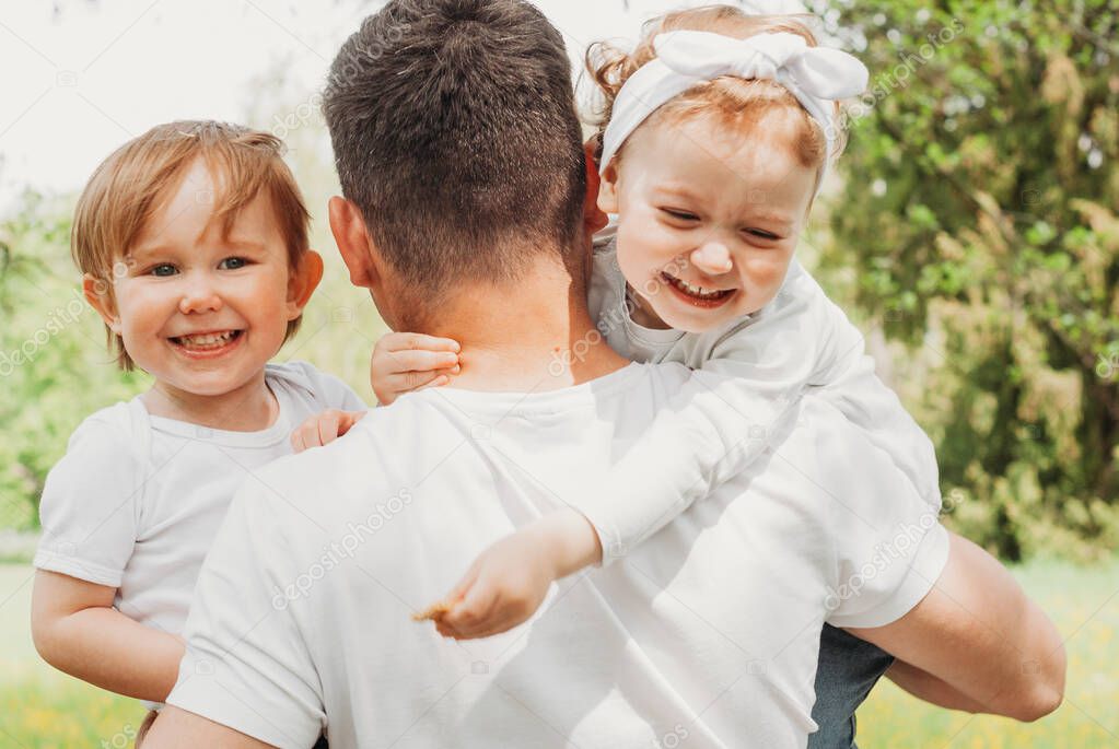 Dad holds two children in his arms. The kids are laughing. Girl hugs dad. Dad and children in white t-shirts. Dad with children walks in the park. A happy family. Father's day