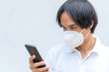 Asyalı bir adam N95 maskesi takıyor ve akıllı telefon kullanıyor. 