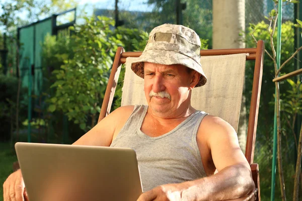 Abuelo sentado en la silla de cubierta en el jardín y mantenga el portátil en sus manos. Concepto de aprendizaje a distancia — Foto de Stock