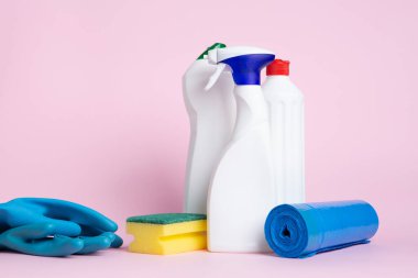 Üç kutu temizlik spreyi, temizleme yastığı, küre ve çöp torbası pembe arka plana yerleştirilmiş.