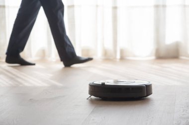 Robot elektrikli süpürge oturma odasını ahşap zeminle temizlerken bir kişi kanepede oturuyor.